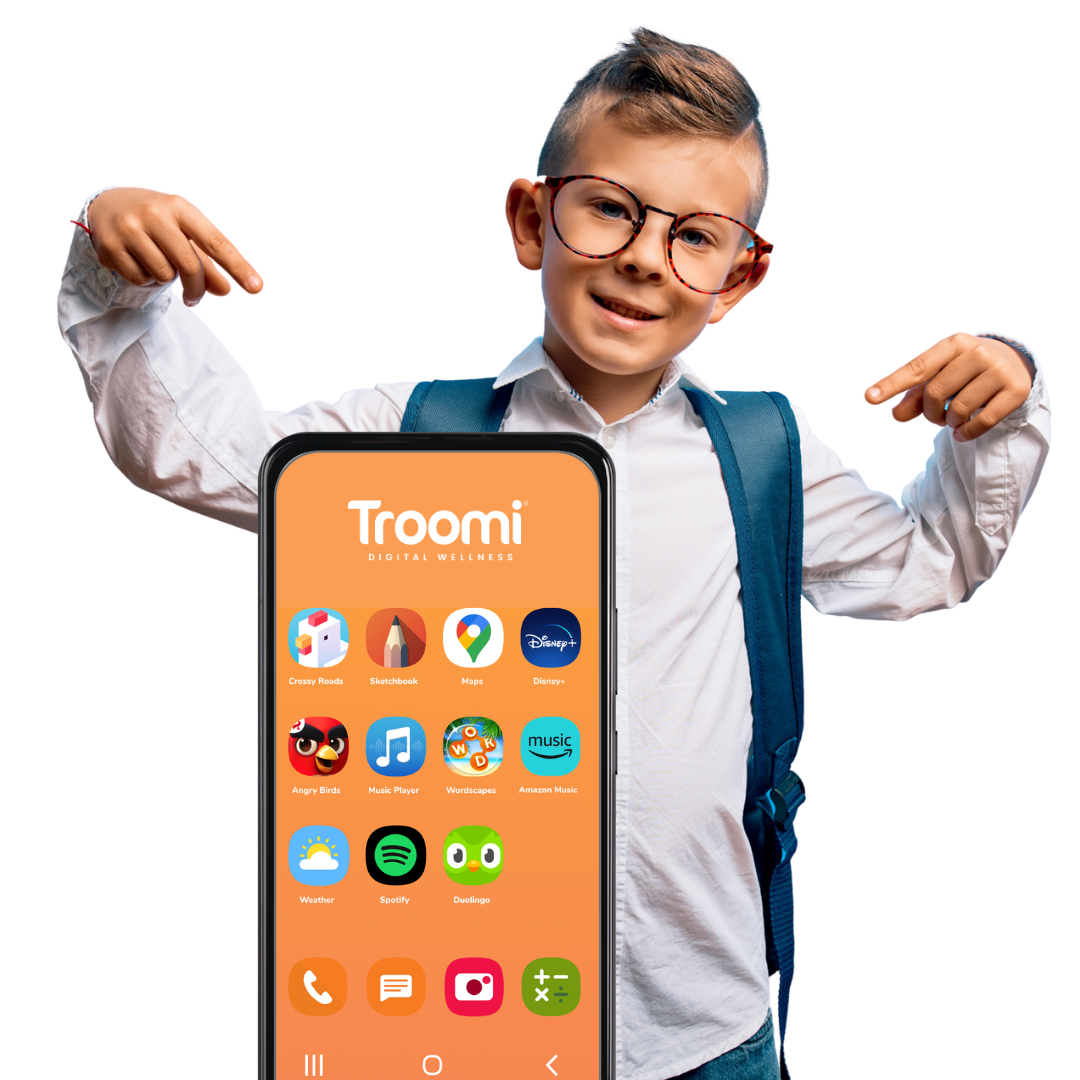A smiling young boy with glasses and a backpack points at a large smartphone displaying the Troomi Digital Wellness home screen, showcasing various app icons on an orange background. Experience safe technology with Troomi.