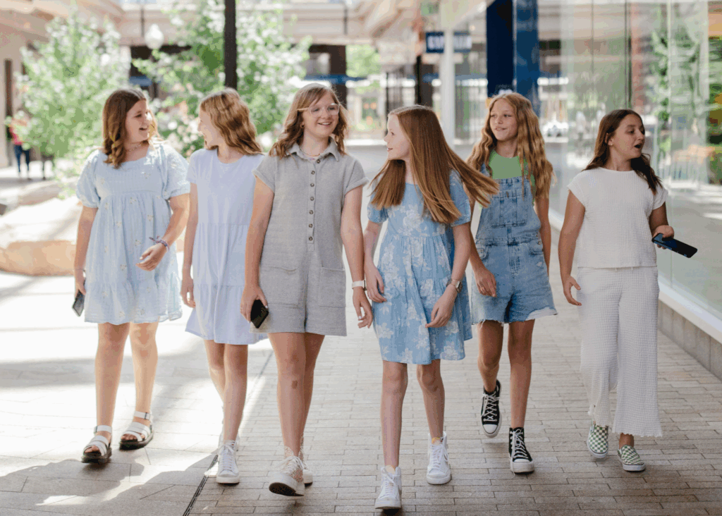Six girls walk together in a shopping area, smiling and talking. They wear casual summer outfits in pastel colors and hold phones. Trees and shop windows line the sunny outdoor walkway.