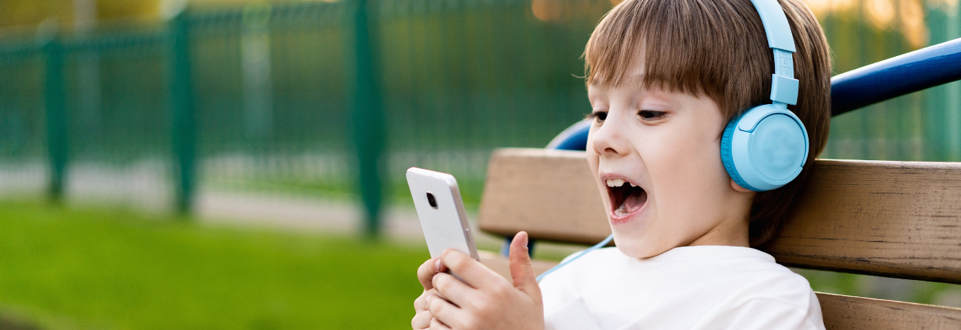A young child sitting on a bench outdoors, wearing bright blue headphones and looking excitedly at a smartphone, with a green fence and grass in the background.