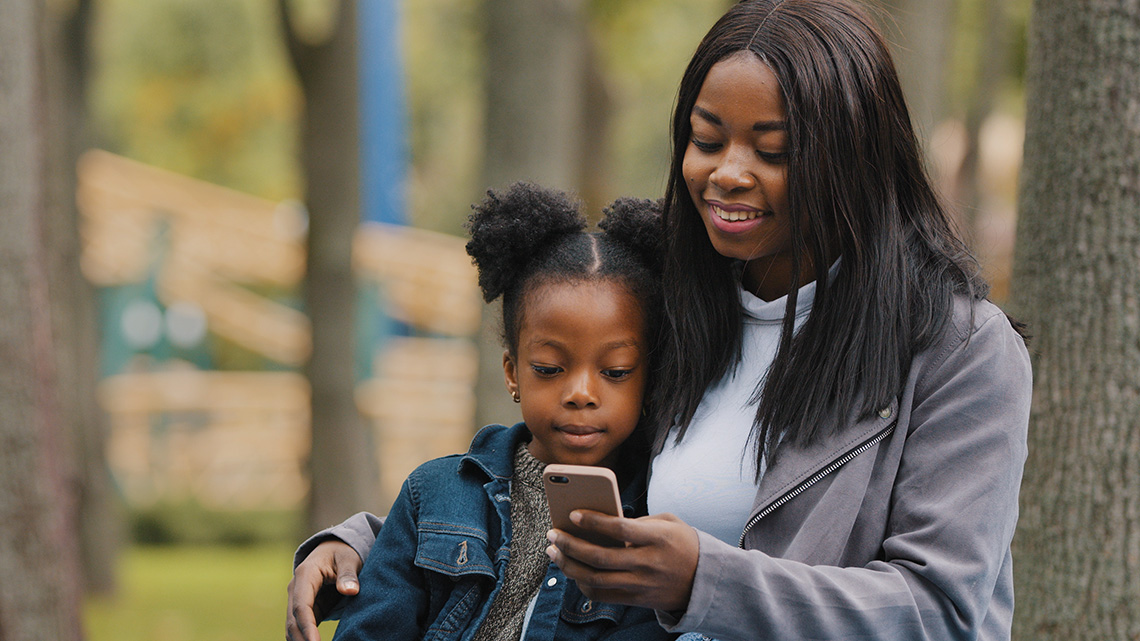 A woman and a young girl sit together outdoors. The woman is smiling and holding a smartphone, while the girl sits close, looking at the phone screen. Trees and blurred background suggest a park setting.