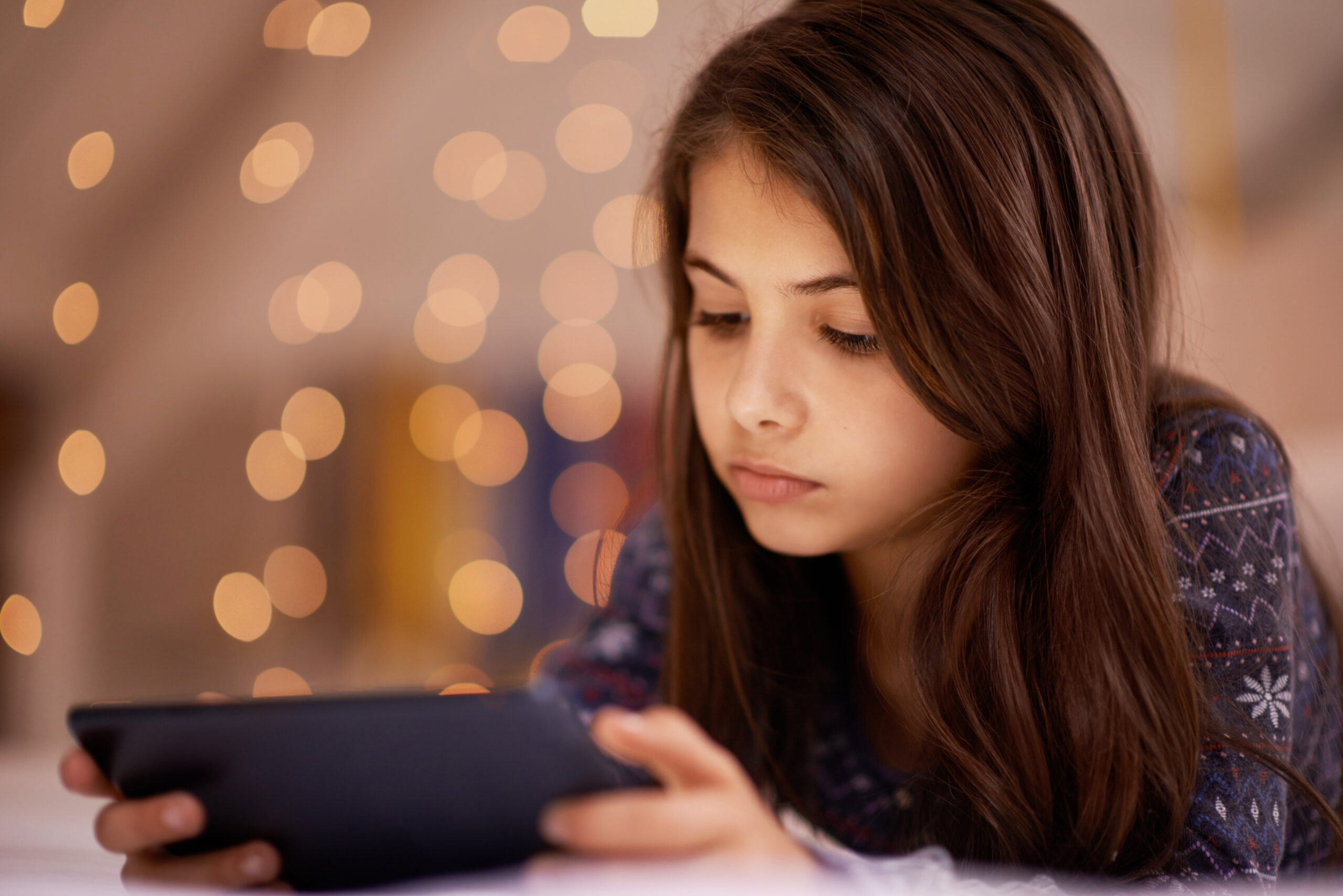 A young girl with long brown hair looks intently at a tablet. She is indoors, with warm, out-of-focus string lights in the background, creating a cozy atmosphere.