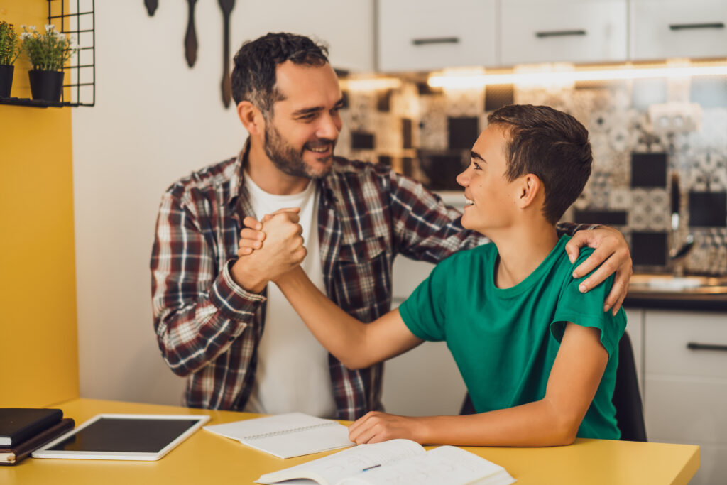 A smiling man and a boy sit at a table in a kitchen, clasping hands in a friendly gesture. Open notebooks and a tablet are on the table, suggesting they are working or studying together.