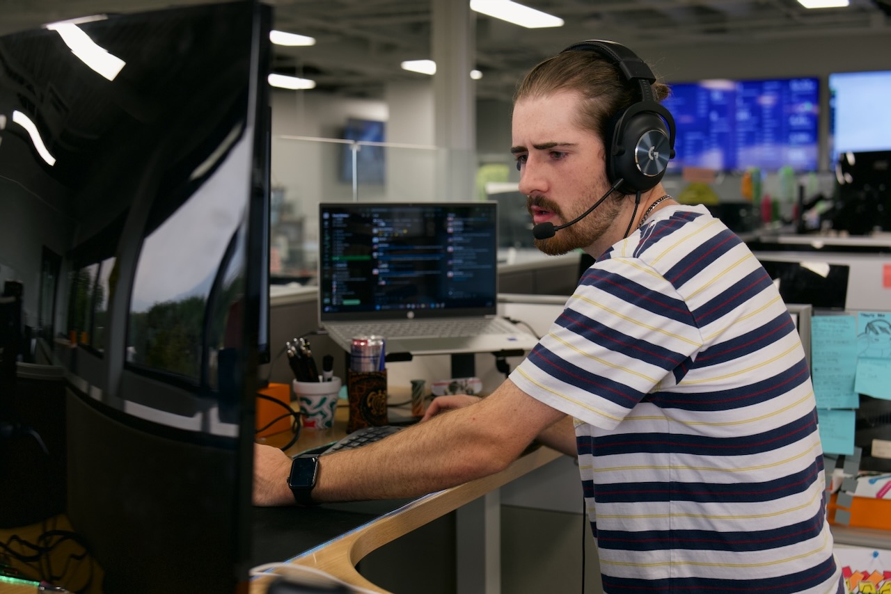 A man wearing a headset and striped shirt sits at a desk with a large monitor, coding on a laptop. He appears focused and is speaking, possibly on a call, in a modern office environment.