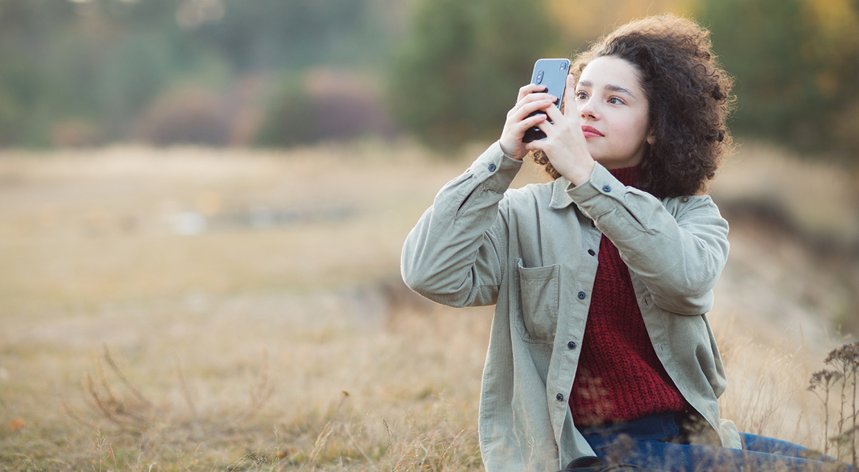 A person with curly hair wearing a green jacket and red sweater holds up a smartphone, taking a photo outdoors in a grassy field with blurred trees in the background.