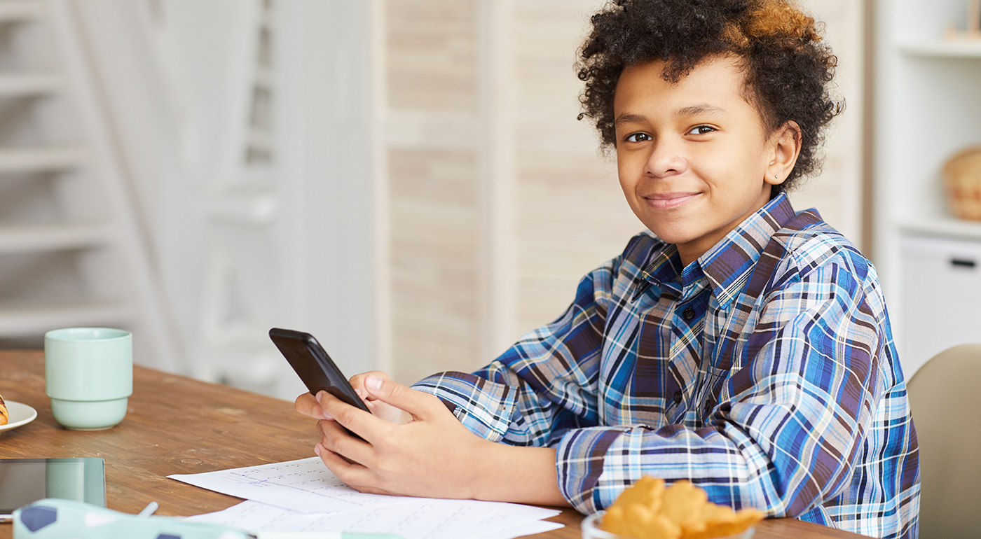 A smiling child with curly hair sits at a table holding a smartphone. There are papers, a croissant, and a light blue mug on the table. The background shows a light-colored room with shelves and stairs.