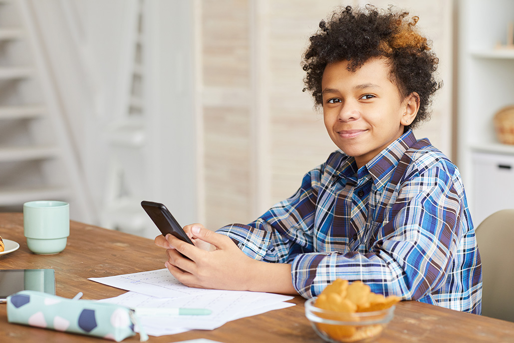A smiling child in a plaid shirt sits at a table with papers, a smartphone, a pencil case, a mug, and a bowl of croissants, looking toward the camera.
