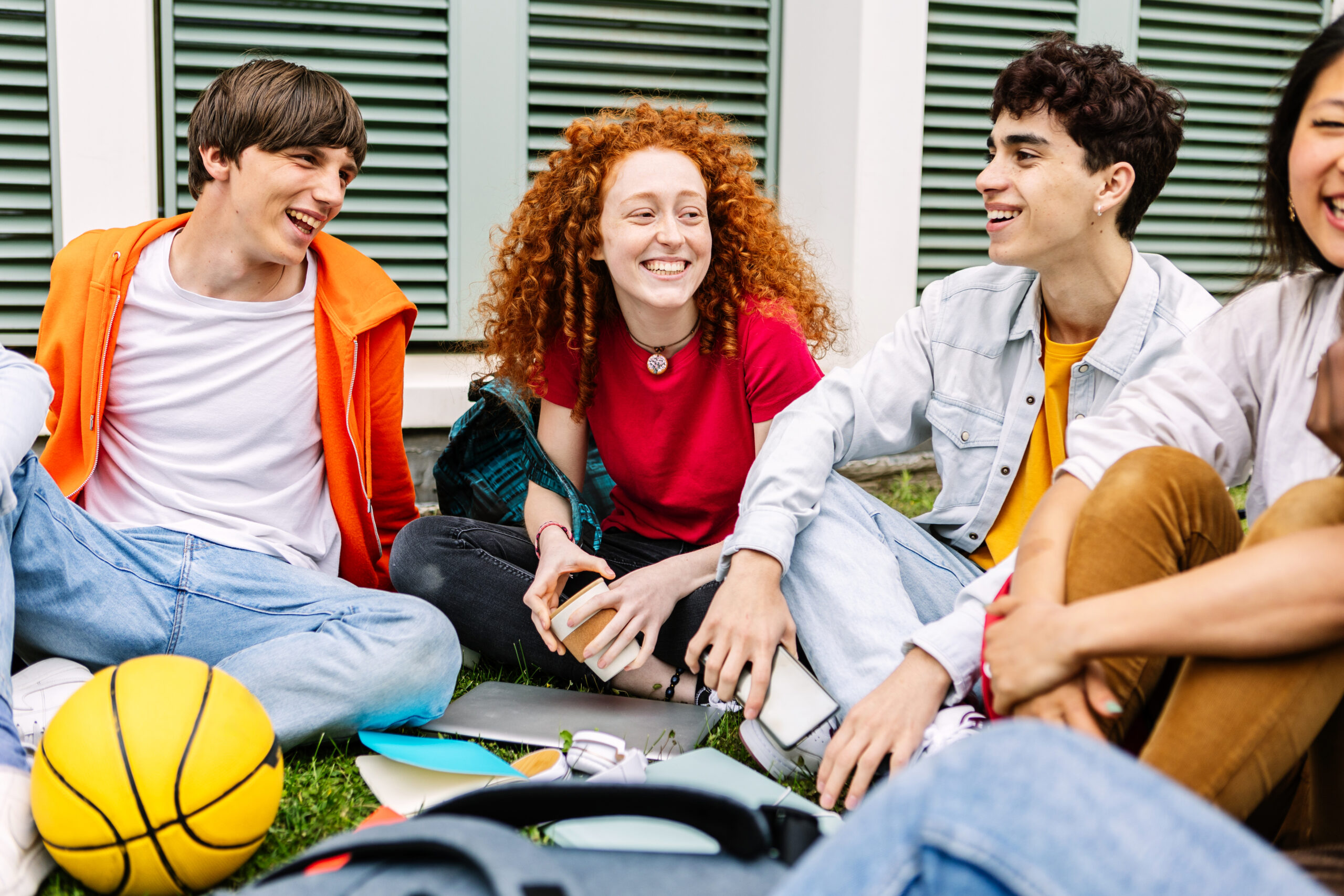 A group of four teenagers sit on grass, smiling and talking together. School supplies and a yellow basketball are nearby. They appear relaxed and happy, enjoying each others company outdoors.