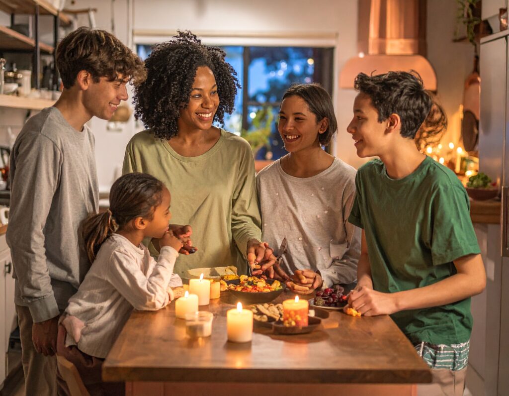 A family of five gathers around a kitchen table, smiling and enjoying food together by candlelight. The warm, cozy setting includes various snacks and glowing candles on the table.