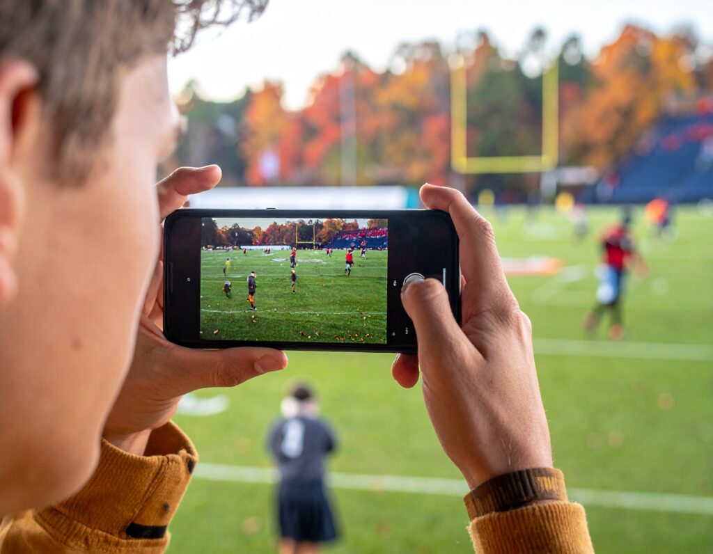A person holds a smartphone and takes a photo of a soccer game on a field. The phone screen shows players and the goalposts, while the background features trees with autumn leaves.