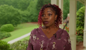 A woman with curly red hair, wearing a mauve floral blouse, speaks outdoors on a porch with greenery and a path visible behind her. She appears to be mid-sentence and is looking slightly off-camera.