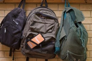 Three backpacks hang on hooks against a beige brick wall. The middle backpack, labeled Vans, has a notebook and an orange pack labeled Roomi visible in its front mesh pocket.