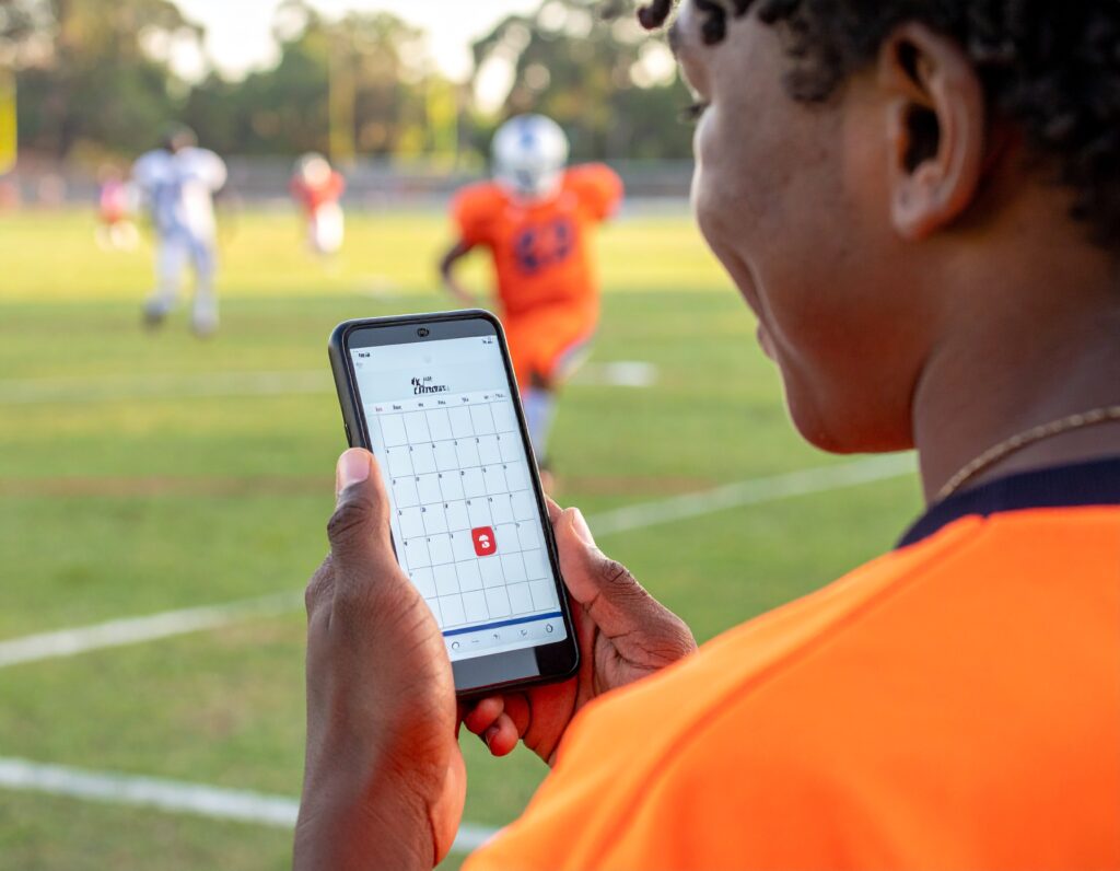 A person in an orange sports jersey looks at a calendar app on a smartphone while standing on a football field, with players in the background during a game or practice.