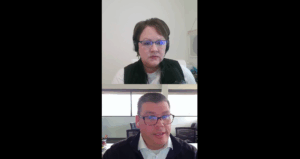 Screenshot of a video call showing two people. The person on top has short brown hair, glasses, and headphones. The person on the bottom has short dark hair, glasses, and is speaking. Both are in office settings.