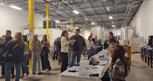 People mingle and talk in a large indoor space with high ceilings and industrial tanks. Several tables with seated women face the camera, covered in white cloths with papers and bags. Bright yellow pillars line the room.