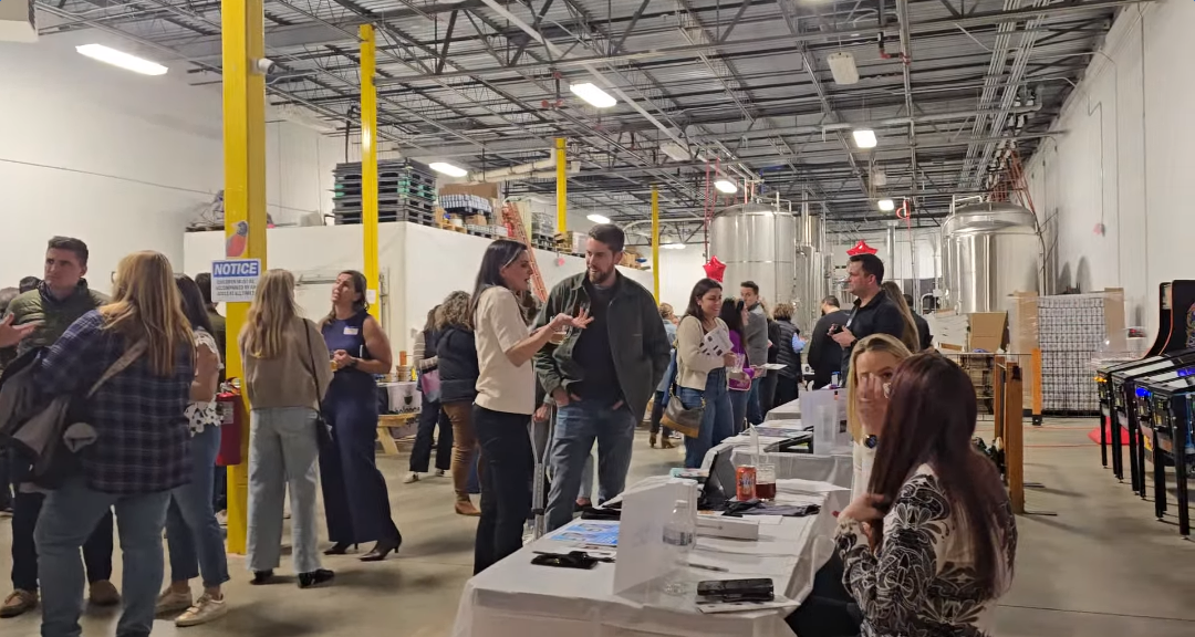 People mingle and talk in a large indoor space with high ceilings and industrial tanks. Several tables with seated women face the camera, covered in white cloths with papers and bags. Bright yellow pillars line the room.