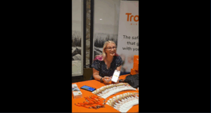 A smiling woman with pink highlights sits at an orange table displaying promotional items, holding up a phone. Behind her is a banner and a black-and-white photo of trees on the wall.