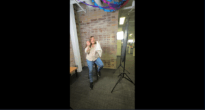 A person stands on one leg, smiling and making a peace sign in front of a brick wall. Photography lights and office decor are visible in the background.