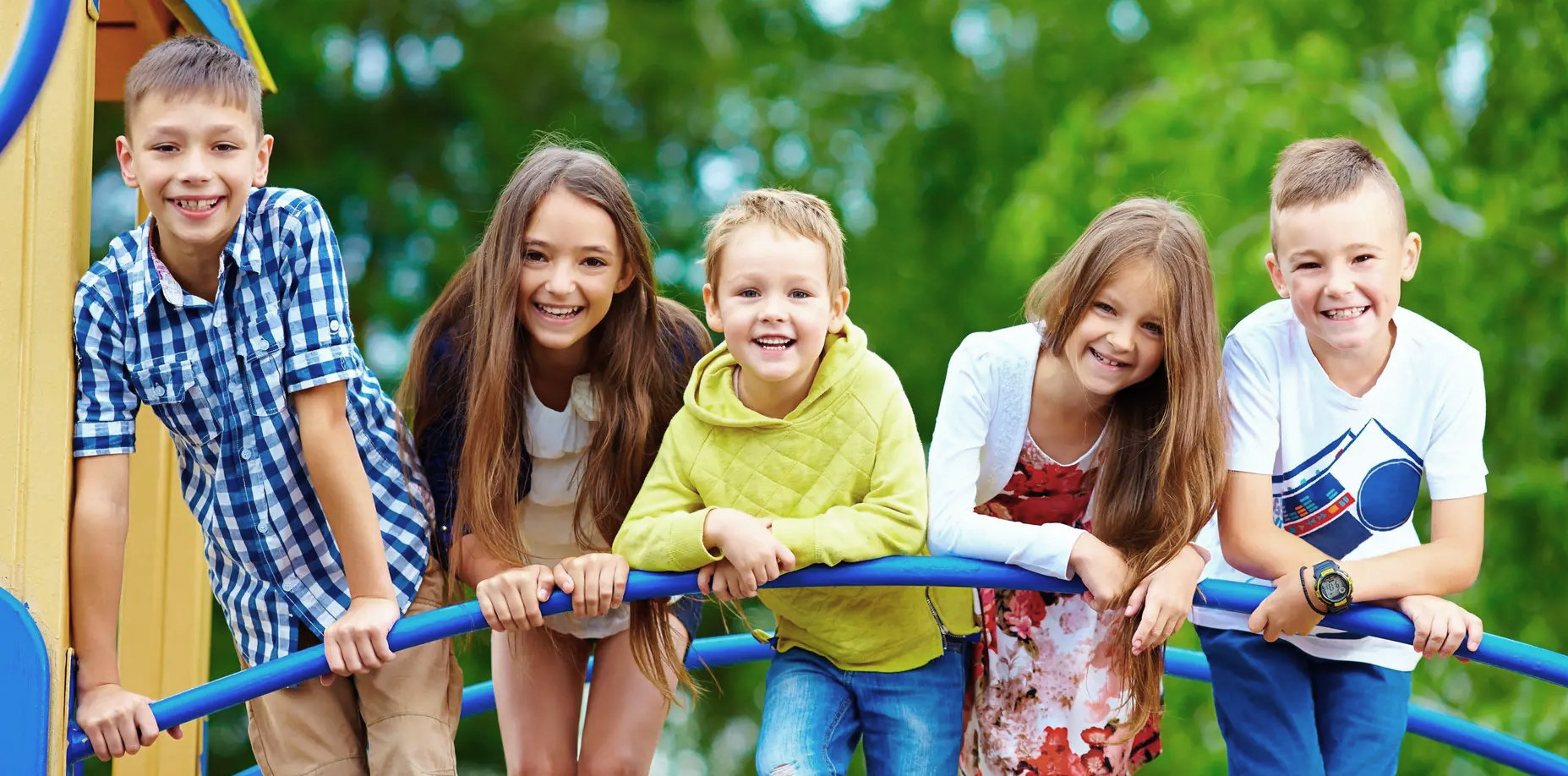 Five smiling children stand outdoors, leaning on blue bars of playground equipment, with green trees blurred in the background. They are casually dressed and appear happy and cheerful.