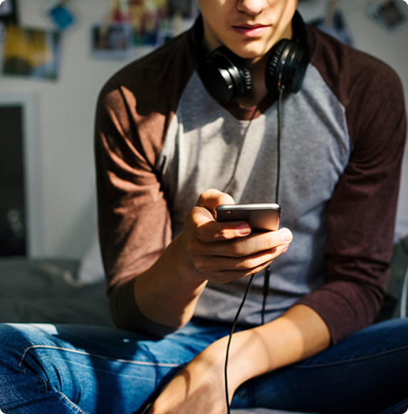 A person wearing a brown and gray long-sleeve shirt and jeans sits cross-legged on a bed, holding a smartphone, with wired headphones resting around their neck. The background is softly out of focus.