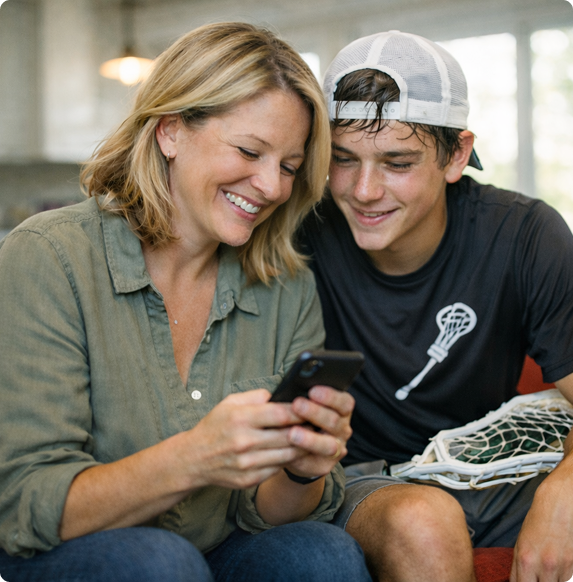 A smiling woman and a teenage boy in a backward cap sit on a couch, looking at a smartphone together. The boy wears a lacrosse shirt and holds a lacrosse stick, suggesting a positive, shared moment.