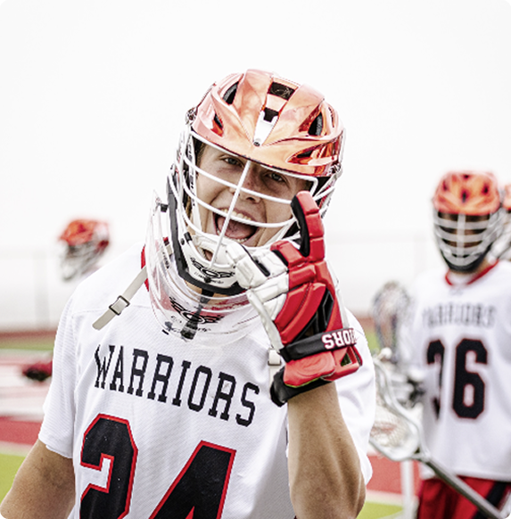 A lacrosse player in a white Warriors jersey and helmet smiles and gestures with his gloved hand toward the camera, while another player stands in the background on the field.