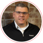 A man with short hair and glasses is smiling, wearing a black jacket with the word Troomi on it, and standing in front of a brick wall.