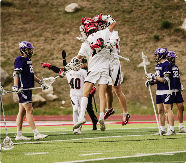 Two lacrosse players in white uniforms jump and hug in celebration on a field, while teammates and opposing players in purple uniforms stand nearby.