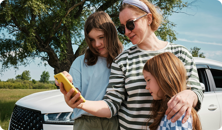 A woman with two young girls stands by a white car outdoors, all looking at a yellow smartphone together. The scene is bright and sunny with green trees and grass in the background.