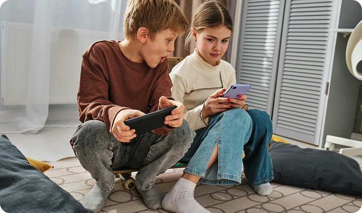 Two children sit together on the floor indoors, each holding a smartphone. The boy on the left looks at the girls phone with surprise, while the girl on the right focuses on her own device. Pillows and a rug are visible around them.
