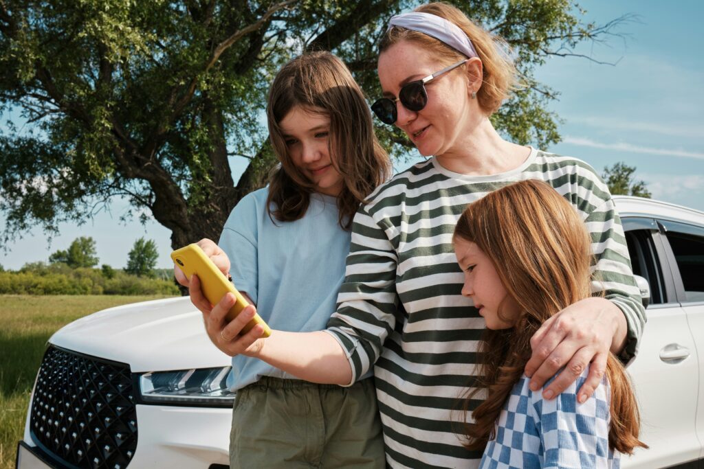 A woman and two girls stand beside a white car outdoors, looking at a yellow smartphone. The woman hugs the girls, and they all appear interested in the phone screen. Trees and grass can be seen in the background.