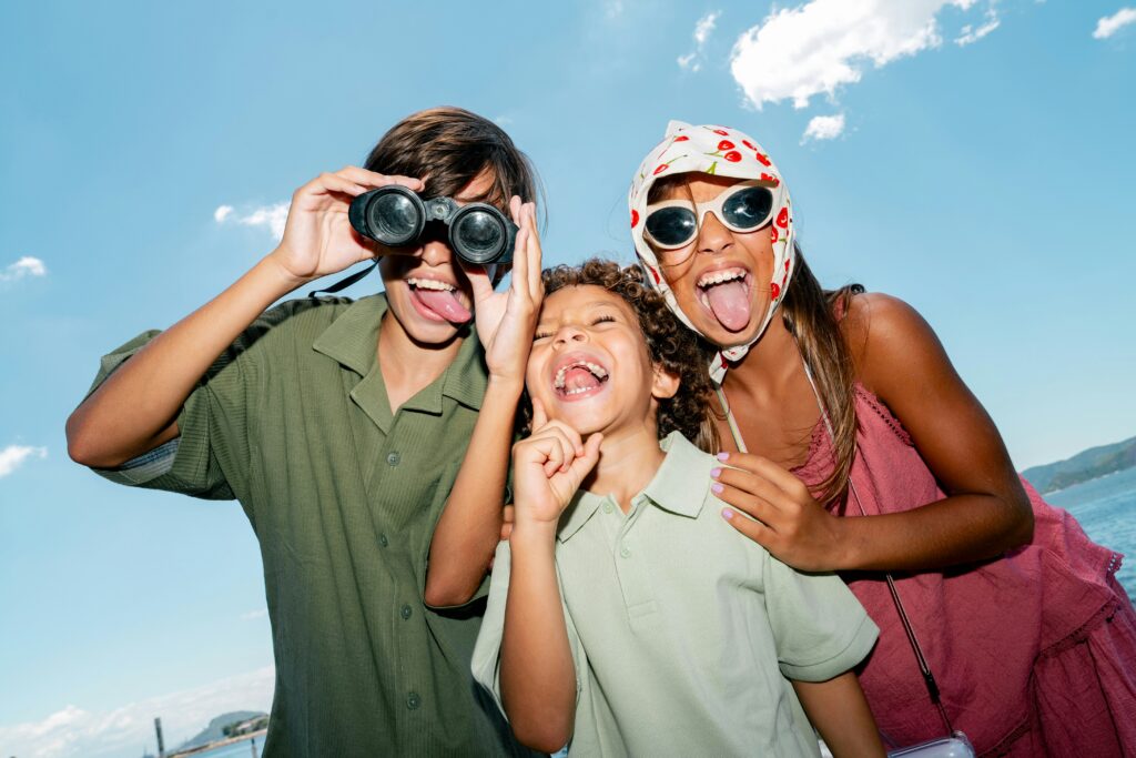 Three children stand outdoors on a sunny day, laughing and sticking out their tongues. One holds binoculars, another points upward, and the third wears sunglasses and a scarf, with water and blue sky in the background.