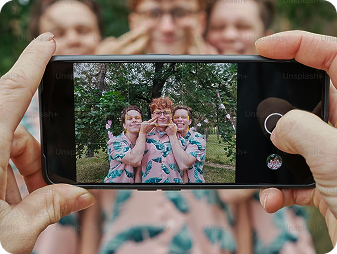 A close-up of hands holding a phone taking a photo of three smiling people in matching outfits, posing outdoors in front of green trees. The group stands close together, appearing happy and playful.