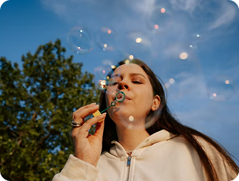 A young woman with long dark hair blows soap bubbles outdoors, wearing a light hoodie. The sky is blue with scattered clouds, and trees are in the background. The bubbles catch the sunlight, creating small reflections.