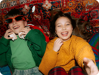 Two kids lying on a colorful patterned rug, laughing. One wears red sunglasses and holds headphones, the other wears a yellow sweater and reaches toward the camera.