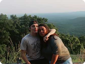 Two people sit close together on a hillside, one with their arm around the other. They are smiling with a scenic, tree-filled valley and distant blue hills in the background. The atmosphere appears relaxed and friendly.