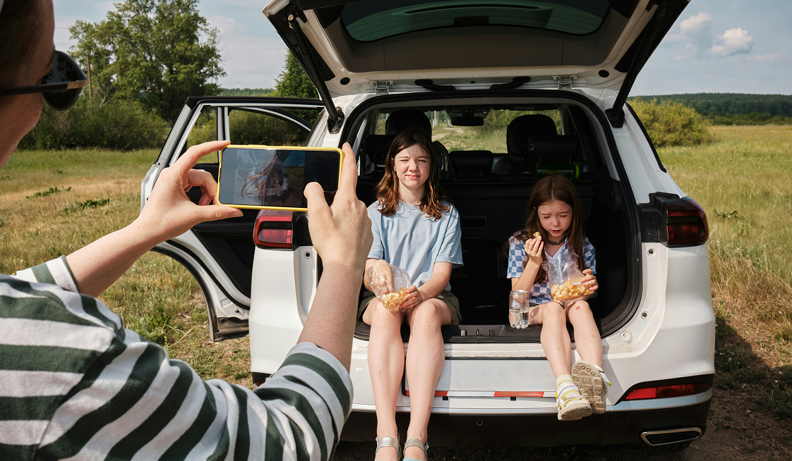 Two children sit on the open trunk of a white SUV, eating snacks, while an adult takes their photo with a smartphone in a grassy, outdoor setting.