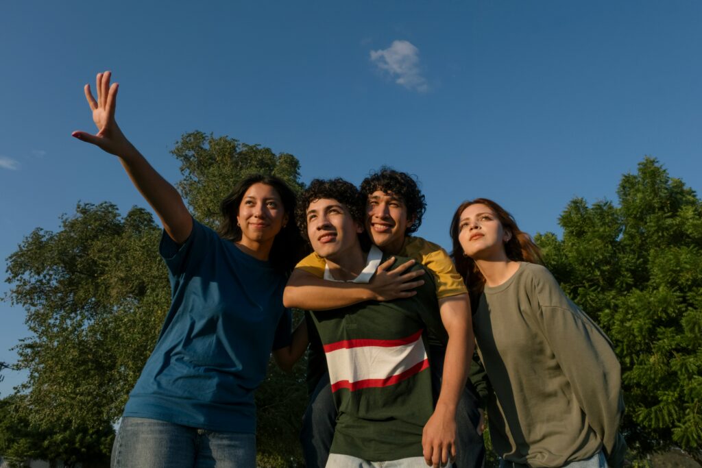 Four young people stand outdoors under a blue sky, smiling and looking into the distance. One person is waving, another is giving a piggyback ride. Green trees are in the background.