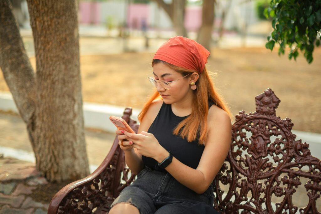A woman with long hair, wearing glasses, a red headscarf, and a black sleeveless top, sits on an ornate outdoor bench looking at her phone. Trees and a blurry background suggest a park setting.