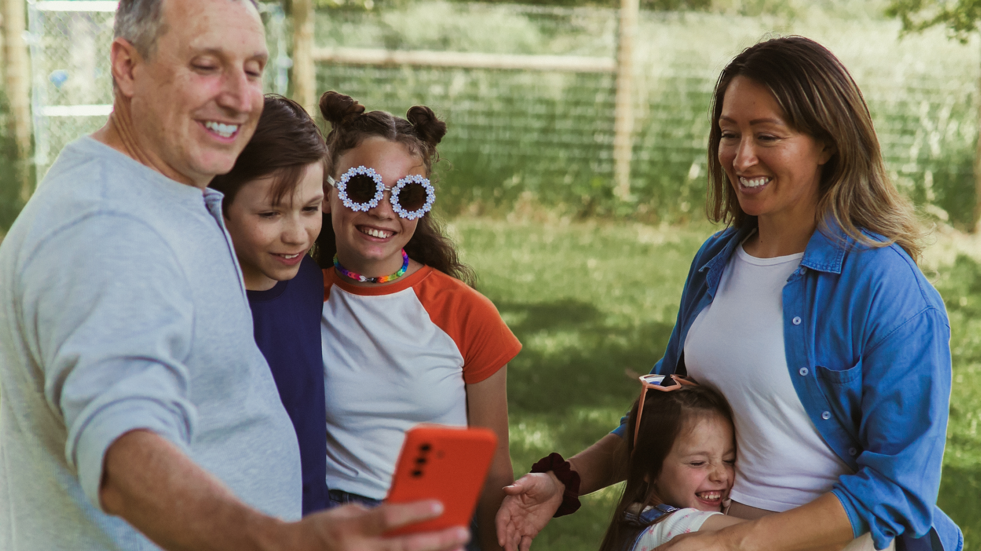 A smiling family stands outdoors, posing for a selfie. An older man holds a phone, with three children and a woman gathered close, one child wearing large daisy-shaped sunglasses. Trees and a fence are visible in the background.
