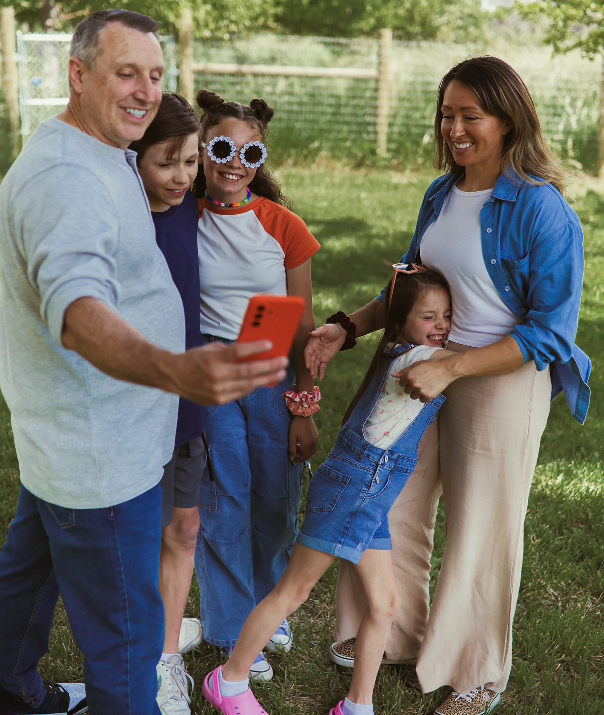 A smiling family of five stands outside on grass, posing for a group selfie. An adult man holds a red phone, while two children and two women, one in playful sunglasses, smile and stand close together. Trees and a fence are in the background.