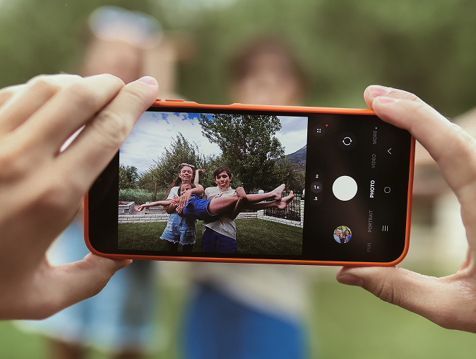 Close-up of hands holding a smartphone, taking a photo of two kids smiling and holding up a third child outdoors with trees and grass in the background.