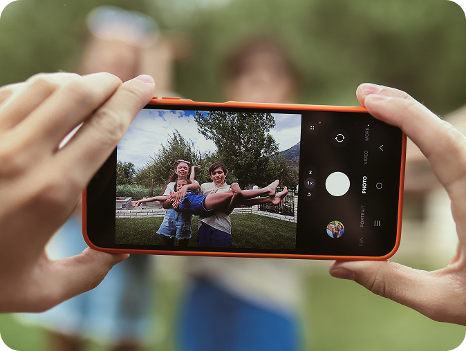 A close-up of a phone capturing two kids outdoors, one holding the other horizontally in their arms, with trees and a fence visible in the background. The scene is in focus on the phone screen.