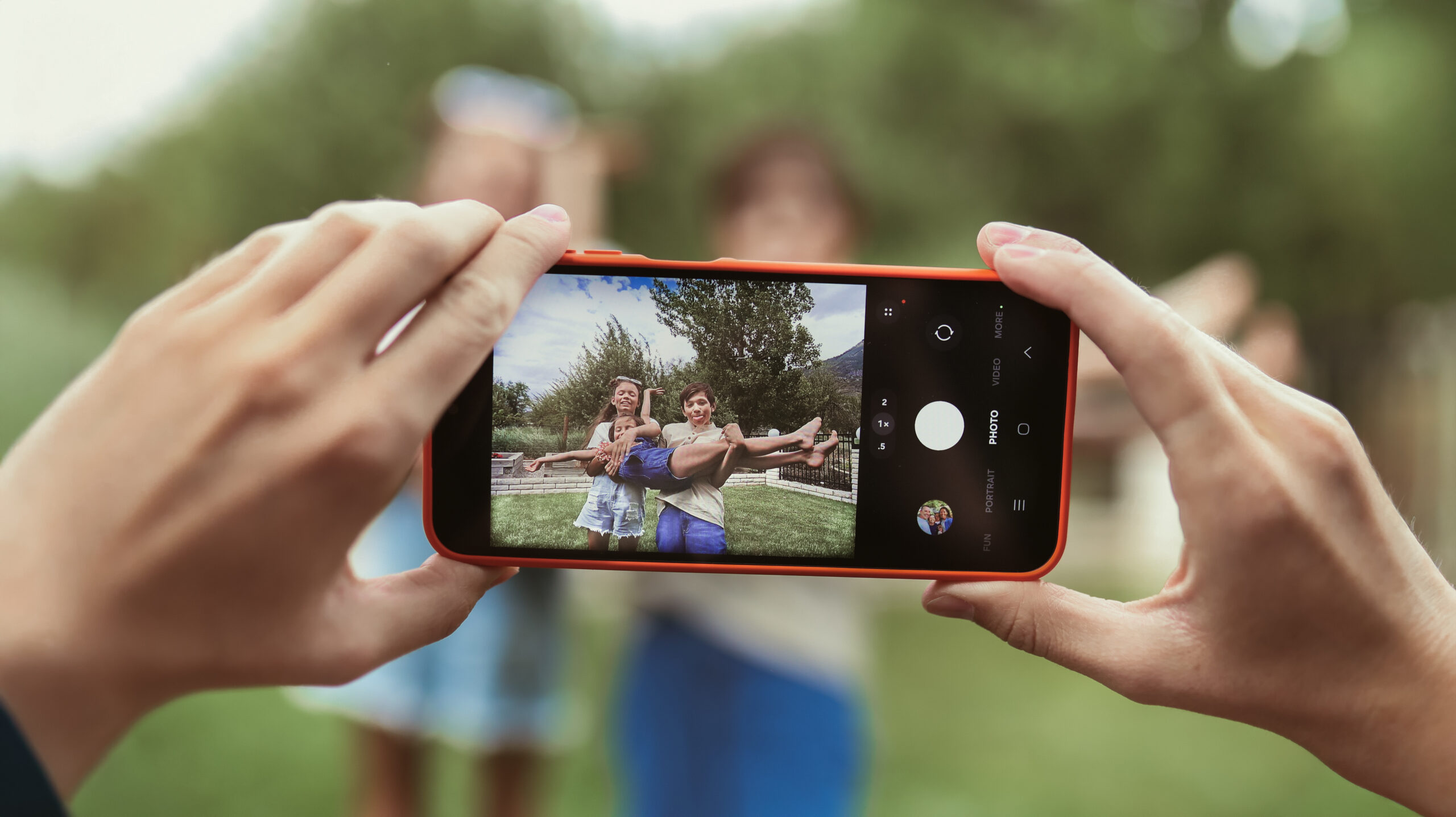 Close-up of hands holding a smartphone taking a photo of two children outside. The kids are posing playfully, with one lifting the other, and greenery is visible in the blurred background.