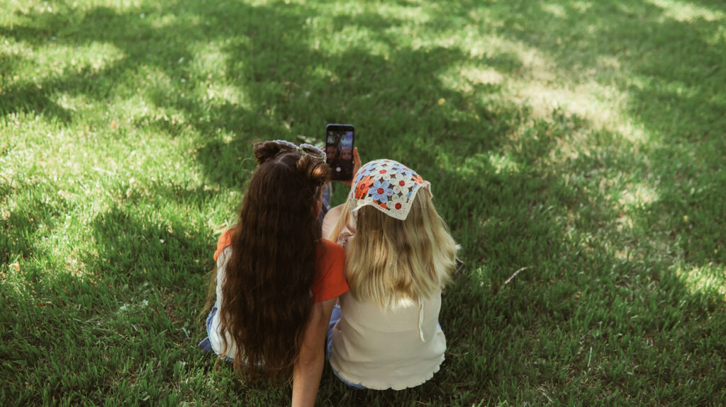 Two girls sit on green grass, seen from behind. One has long brown hair, the other wears a floral headscarf. They are close together, looking at a smartphone and taking a selfie. Sunlight creates dappled shadows on the lawn.