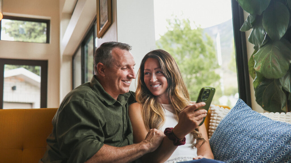 A man and a woman sit on a yellow couch by large windows, smiling and looking at a smartphone together. The scene is bright and cheerful, with plants and pillows adding a cozy touch.