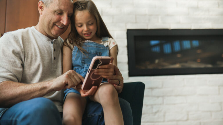 An older man and a young girl sit together, smiling as they look at a smartphone. The girl is wearing denim overalls, and they appear to be enjoying a moment together in a bright, cozy room.