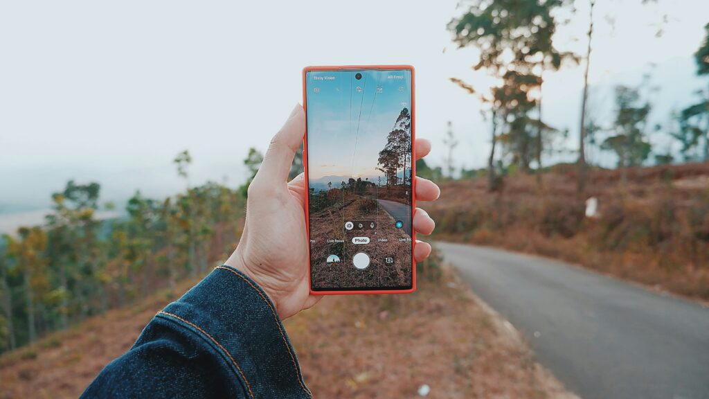 A person holds a smartphone in their left hand, taking a photo of a rural outdoor scene with trees and a curving road; the phone screen displays the landscape being photographed.