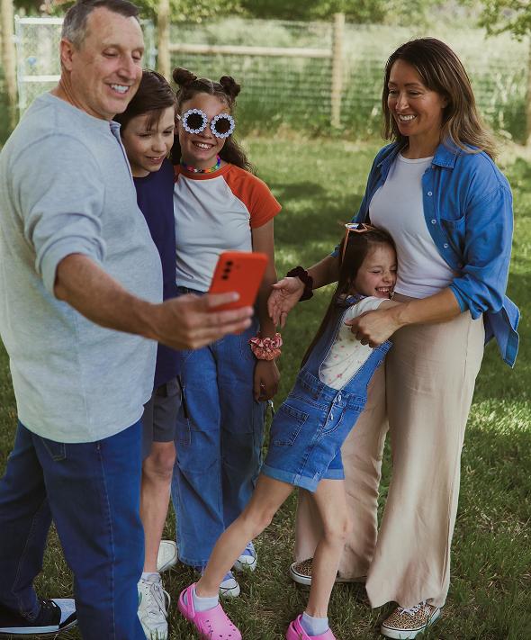 A smiling family of five poses for a selfie outside on grass, with trees and a fence in the background. The man in front holds a red phone, and everyone looks happy and relaxed.