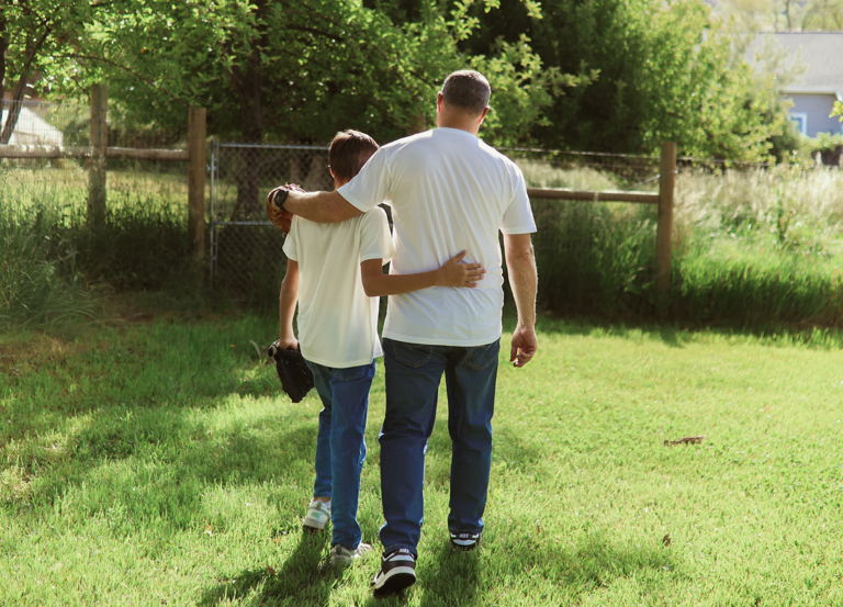 An adult and a child walk together on grass, arms around each other, both wearing white shirts and jeans. The child carries a baseball glove. Trees and a wooden fence are in the background.