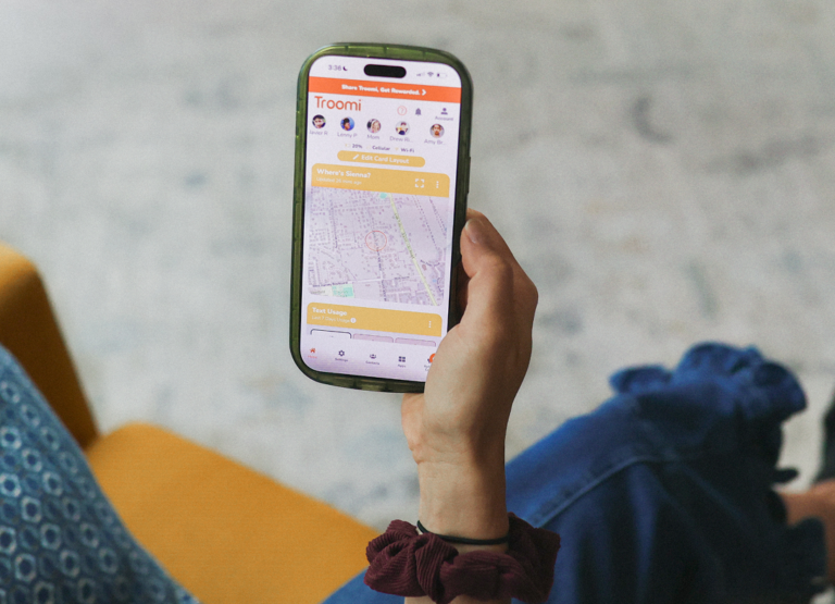 A person holds a smartphone showing a location-tracking app called Troomi, displaying a map with tracked locations and user profiles at the top. The person wears a maroon scrunchie and sits on a yellow chair.